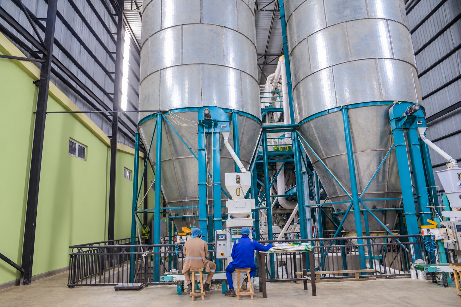 Two operators working at control stations in front of large corn storage silos